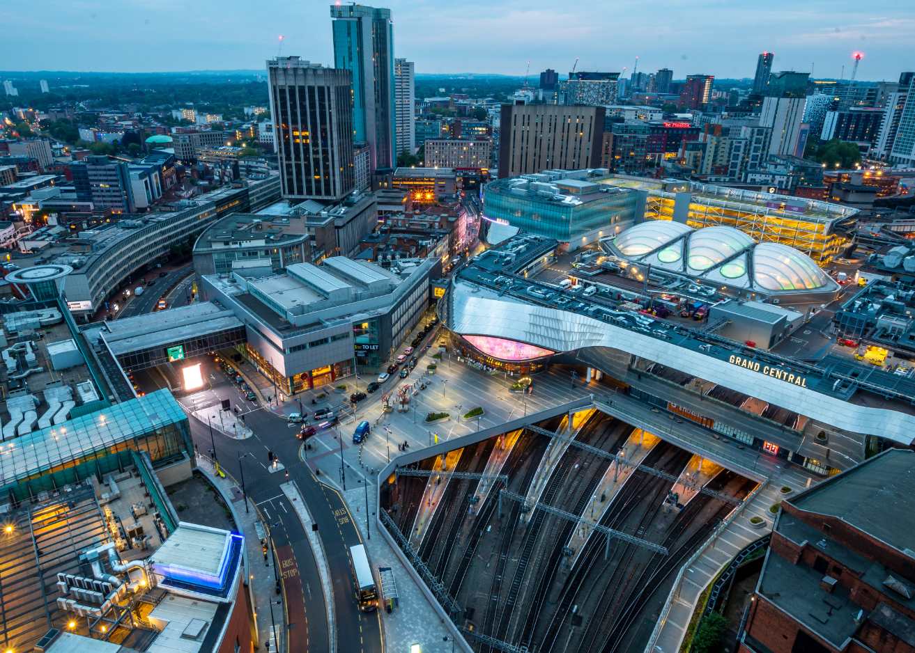 Aerial view of Manchester city at dusk, showcasing illuminated buildings and a vibrant skyline against a twilight sky.