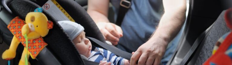 A baby seated in a car seat, happily playing with a colorful toy.