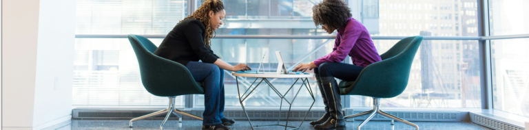 Two women engaged in work at a table, each using a laptop, focused on their tasks in a collaborative environment.