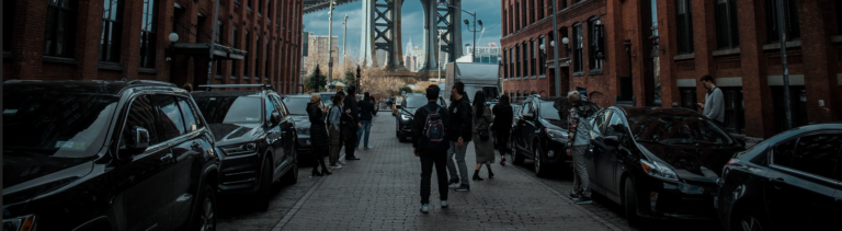 A diverse group of individuals strolling along a street, with a bridge prominently visible in the background.