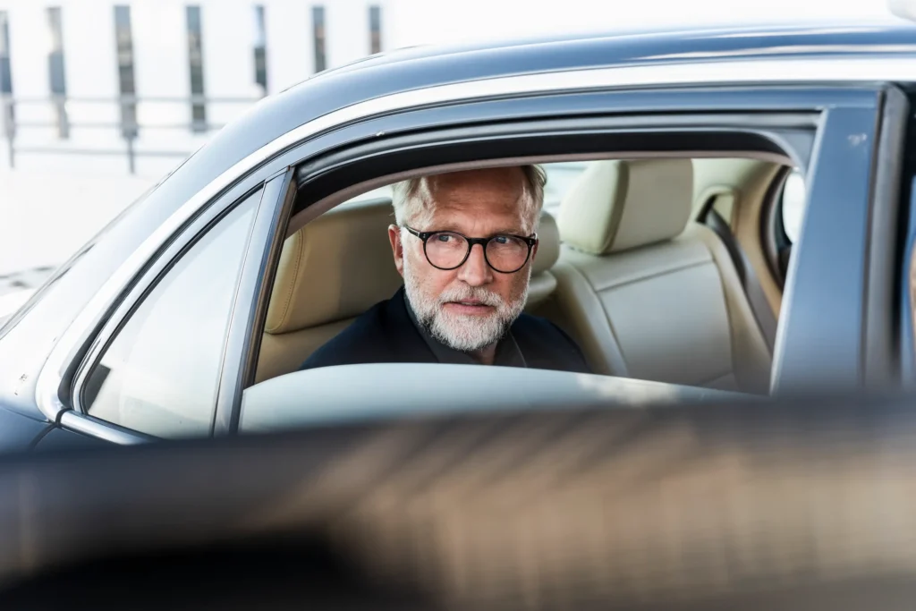 An older man with glasses and a beard is focused on driving a car.