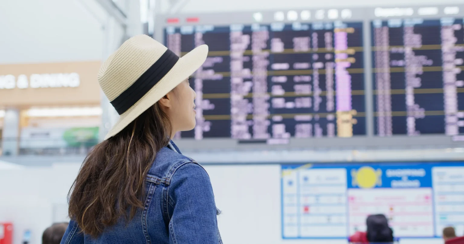 A woman in a hat gazes at an airport departure board, contemplating her travel plans.