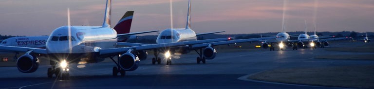 A group of airplanes lined up on the runway during dusk, with a colorful sky in the background.