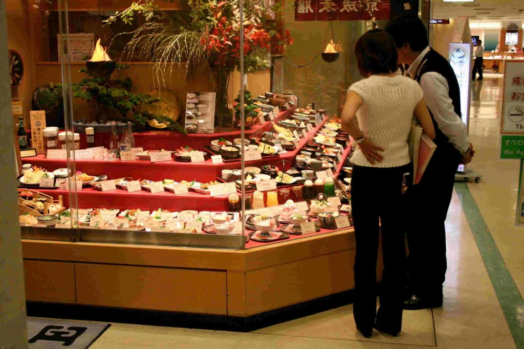 Travelers looking at a vibrant Japanese restaurant food display case while shopping at an airport terminal.
