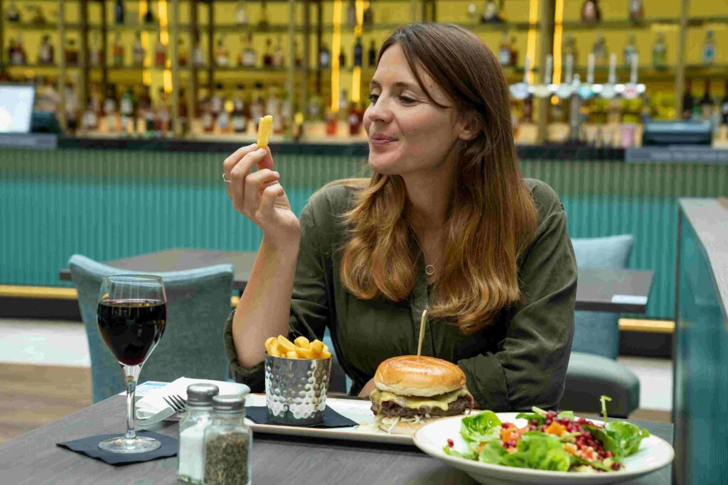 A woman enjoying a burger, fries, salad, and red wine at a modern London airport terminal restaurant with a bar background.