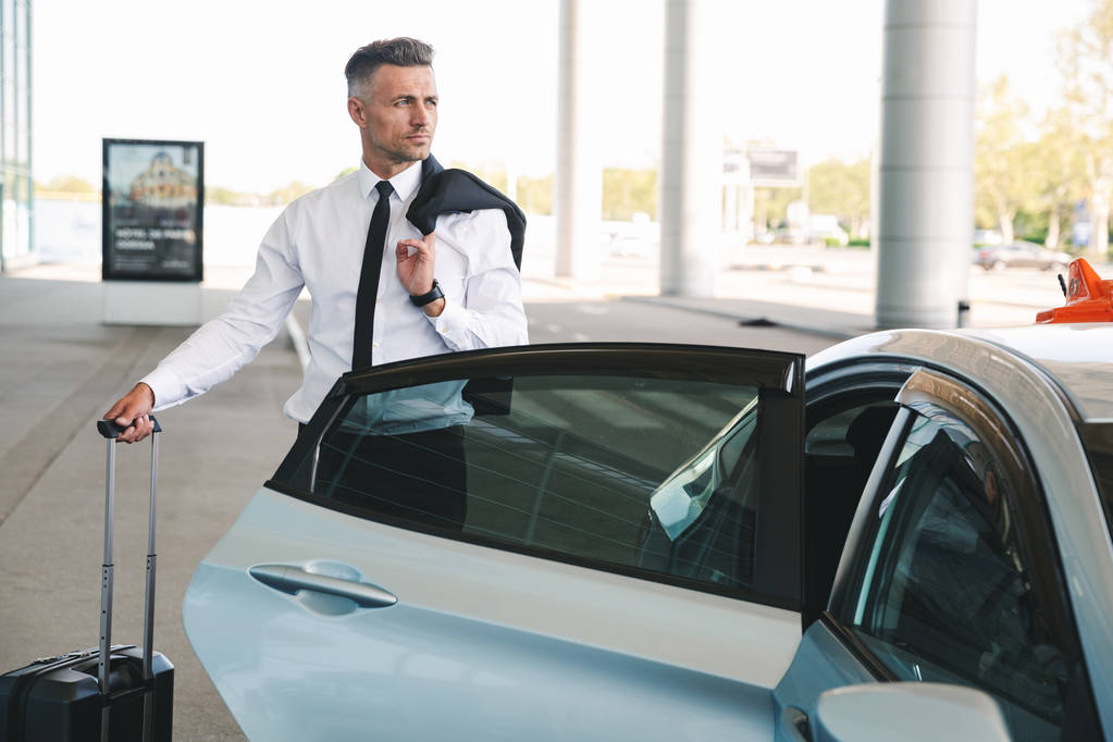 Serious mature businessman getting in taxi outside the airport with a suitcase