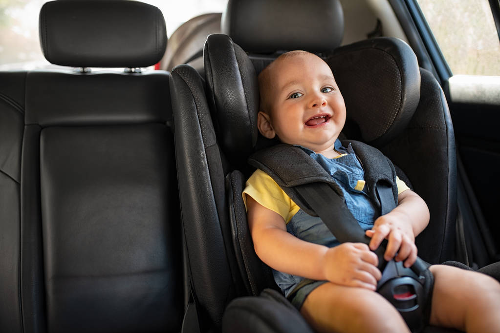 Portrait of happy little child sitting in car seat with safety belt, enjoying road trip. Cute baby boy smiling and having fun while being in the infant car seat. Laughing curious toddler enjoying travel, copy space.