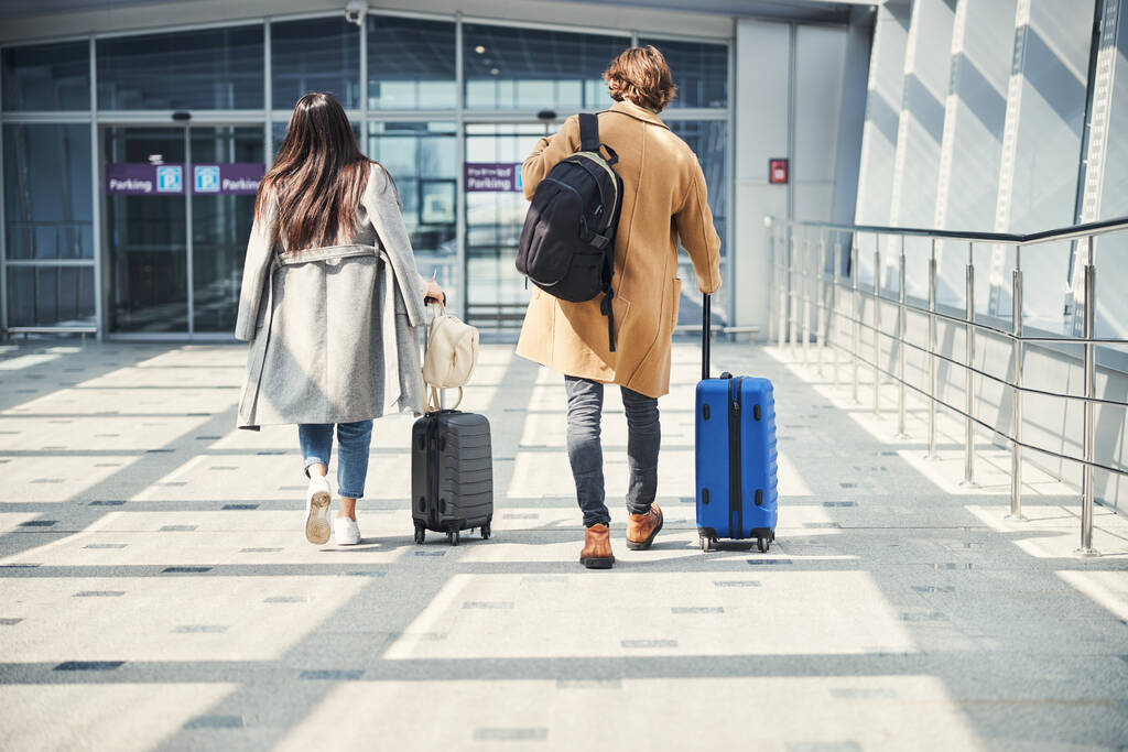 Back view of elegant lady and gentleman with luggage trolley bags heading to airport terminal