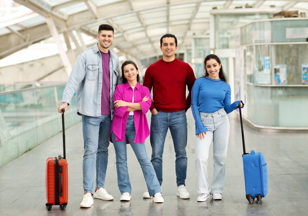 Suitcase Squad. Two Happy Couples Friends Hugging Posing With Their Luggage Ready For Vacation Trip Standing In Modern Airport Indoor, Looking At Camera. Travel Offer Ad. Full Length Shot