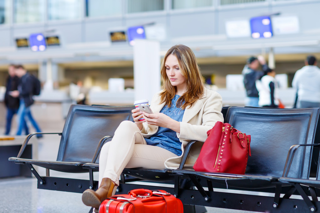 Beautiful girl at international airport, reading her ebook computer and drinking coffee to go while waiting for her flight. Female passenger at terminal, indoors. Traveling people