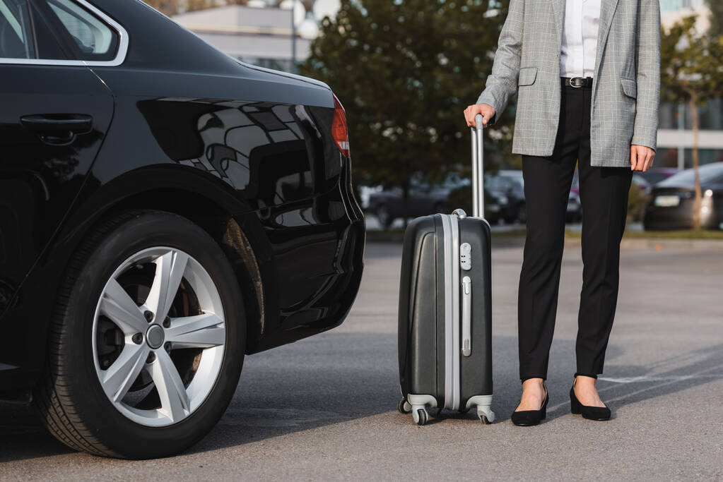 Cropped view of businesswoman holding suitcase near black car
