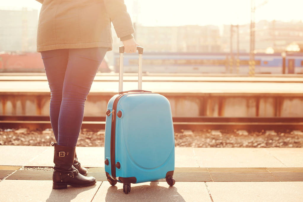 Traveler woman waiting at railway station. Travel by train. Girl standing with blue luggage suitcase in 