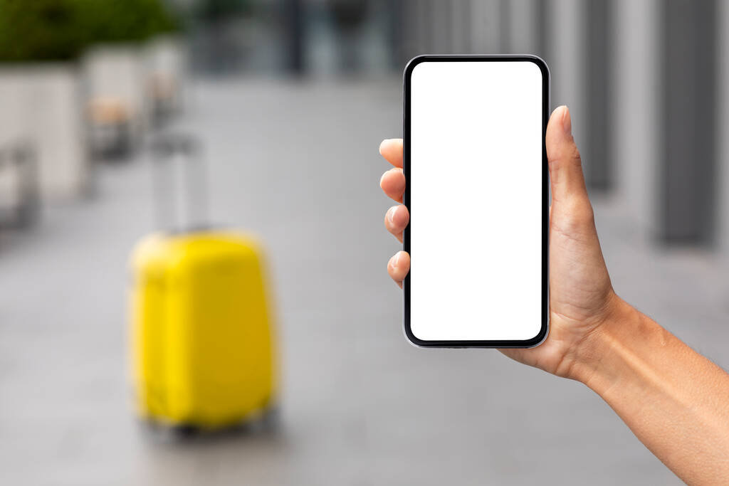 Female hand holding modern smartphone with white blank screen over yellow suitcase, mockup, unrecognizable woman tourist showing nice mobile app for online hotel and flight reservation, copy space