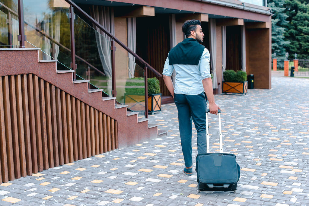 Arabic Muslim Young Man Coming Out of Corner and Goes With Travel Bag to Modern Restaurant, Stopped and Smiling, Holding Black Suitcase. Handsome Muscular Guy Dressed in Black Classic Trousers, White Shirt on Top of Which Knotted Black Sweater. Concept of Business Meeting, Airport, Flight Has Been Delayed, Decides to Explore New Places.