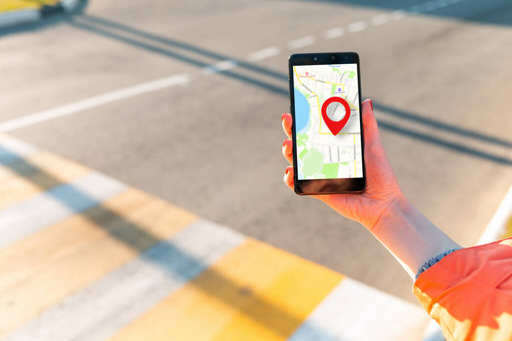 A woman's hand holds a smartphone with an online map that has a red geolocation icon. In the background, a pedestrian crossing in a blur. Copy space. Concept of online navigation and GPS.