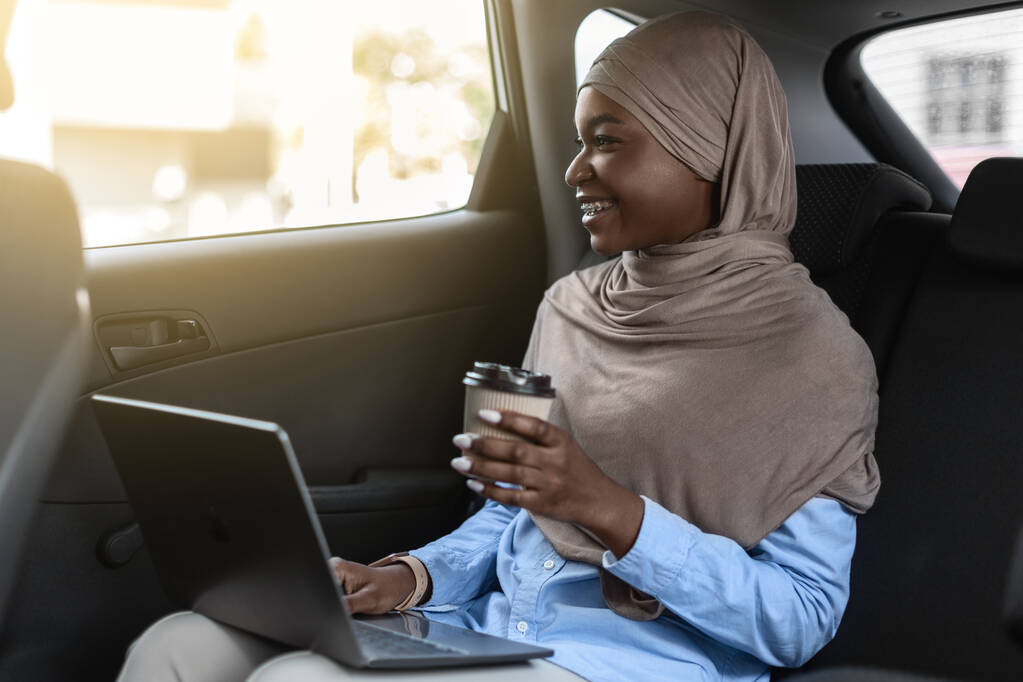 Black muslim businesswoman in hijab riding to office with laptop and coffee on backseat, african islamic lady in headscarf enjoying road ride, having private transfer or using taxi service, free space