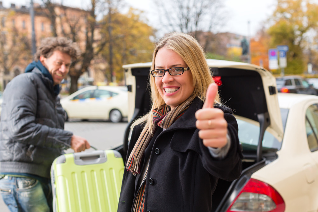 Young woman standing in front of taxi, she has reached her destination, the taxi driver will help with the luggage