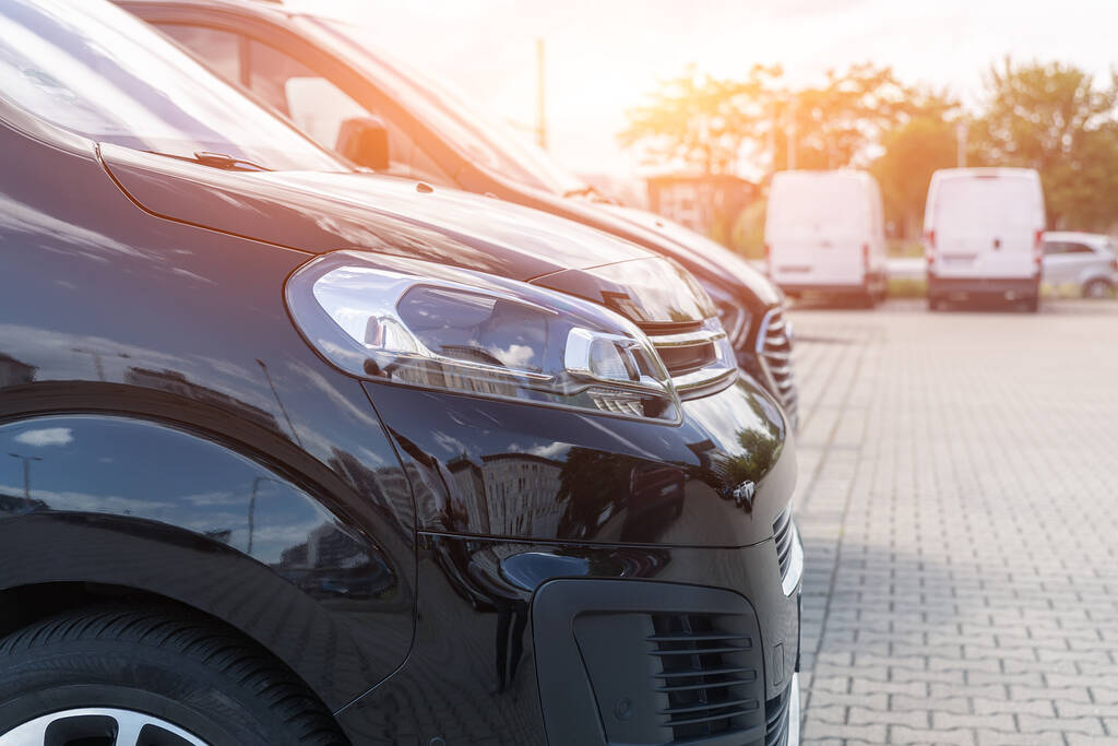 Close-up detail headlight view of many modern luxury black vans parked in row at car sale rental leasing dealer against sunset. Commericial trasfer cargo transportation company fleet. Vip charter.